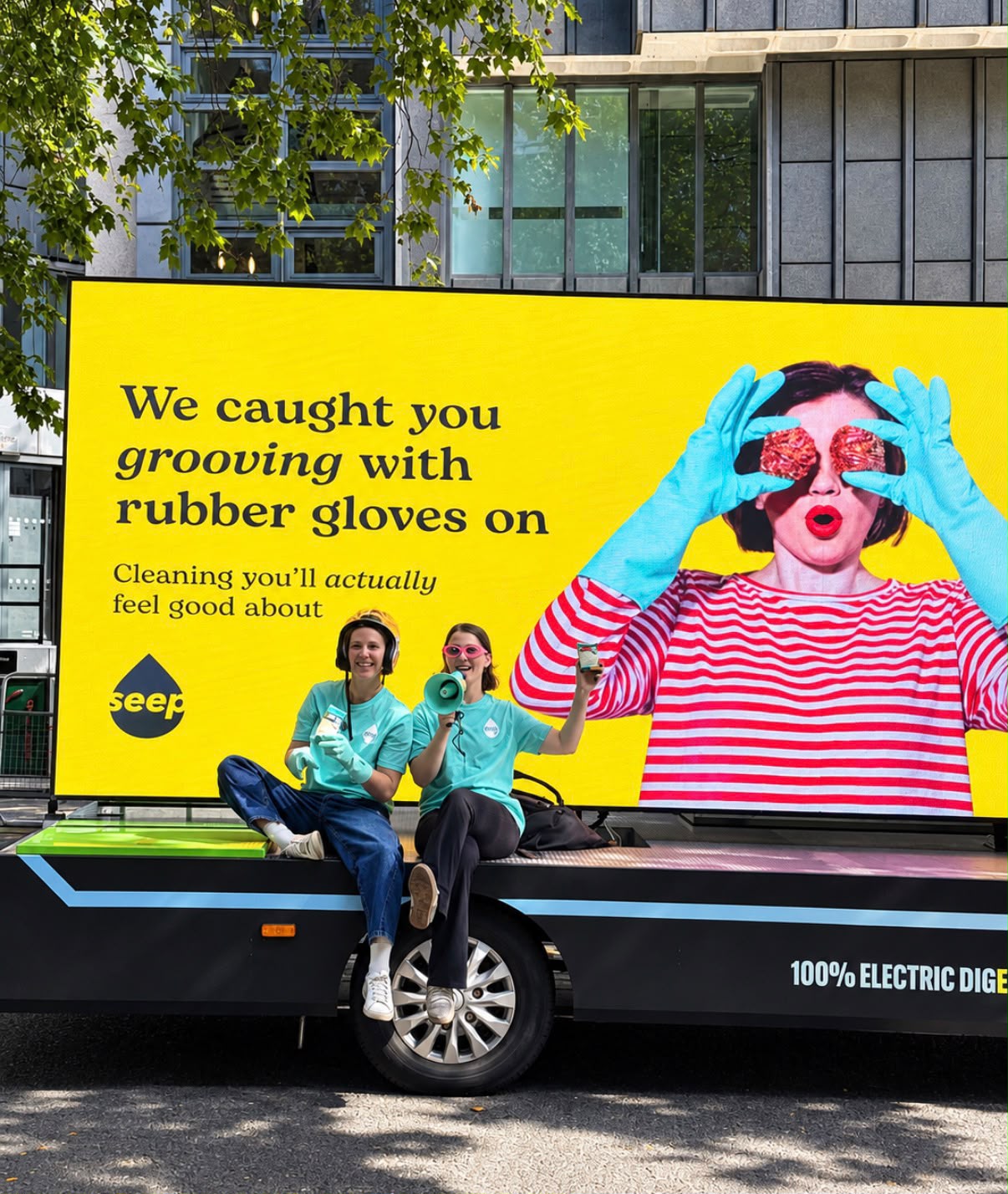 Seep electric Digivan parked outside a glass-fronted building with its screen showing the campaign line 'We caught you grooving with rubber gloves on', and Seep brand ambassadors in teal t-shirts handing out samples